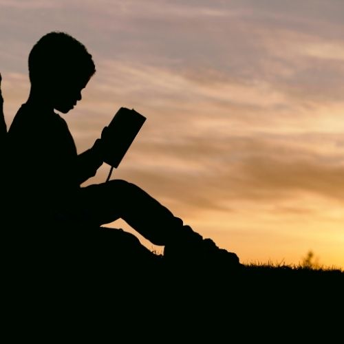 Image of shiloutette of a child reading a book against a tree while the sunsets