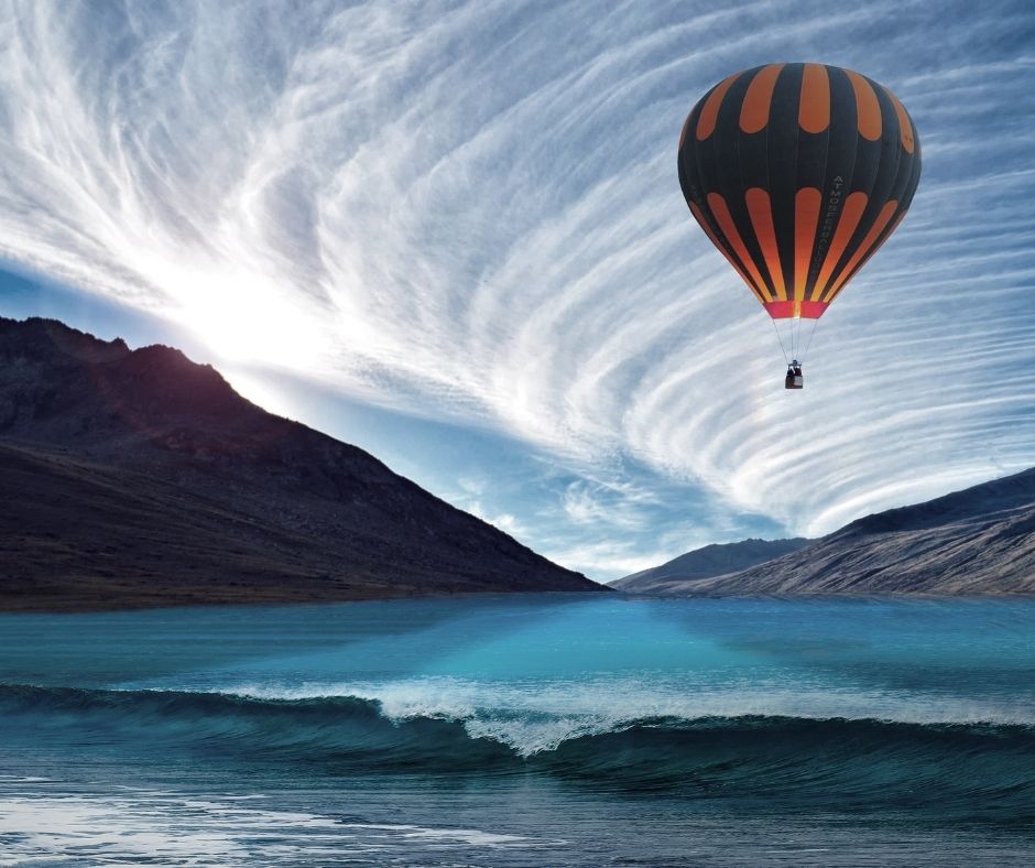 Image of a yellow and blue hot air balloon flying over water with a mountain in the backgrounds. the clouds arch up behind it looking almost like a crashing wave