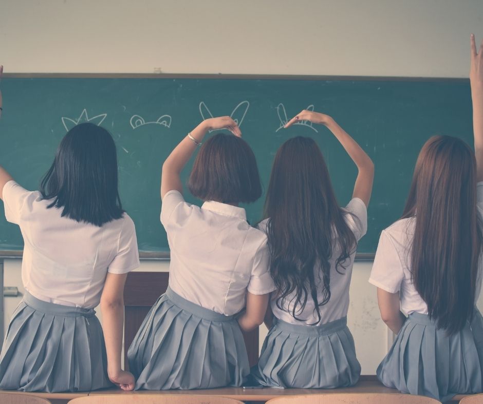 image of four girls in school uniforms sitting with their backs to the cameras facing a chalkboard. the second girl from the left has her left arm raised and curved over her head. the third girl from the left has her right hand raised and curved over her head. the two sort of make a heart with their arms?
