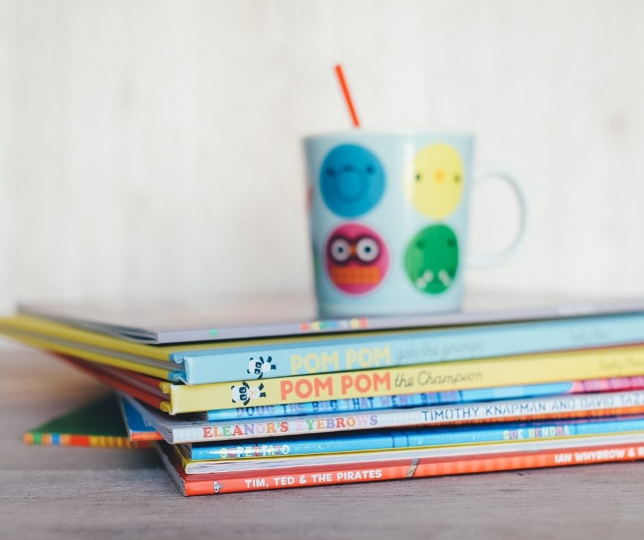 Image description: a stack of colorful children's books are stacked spine out with a brightly colored coffee mug resting on top