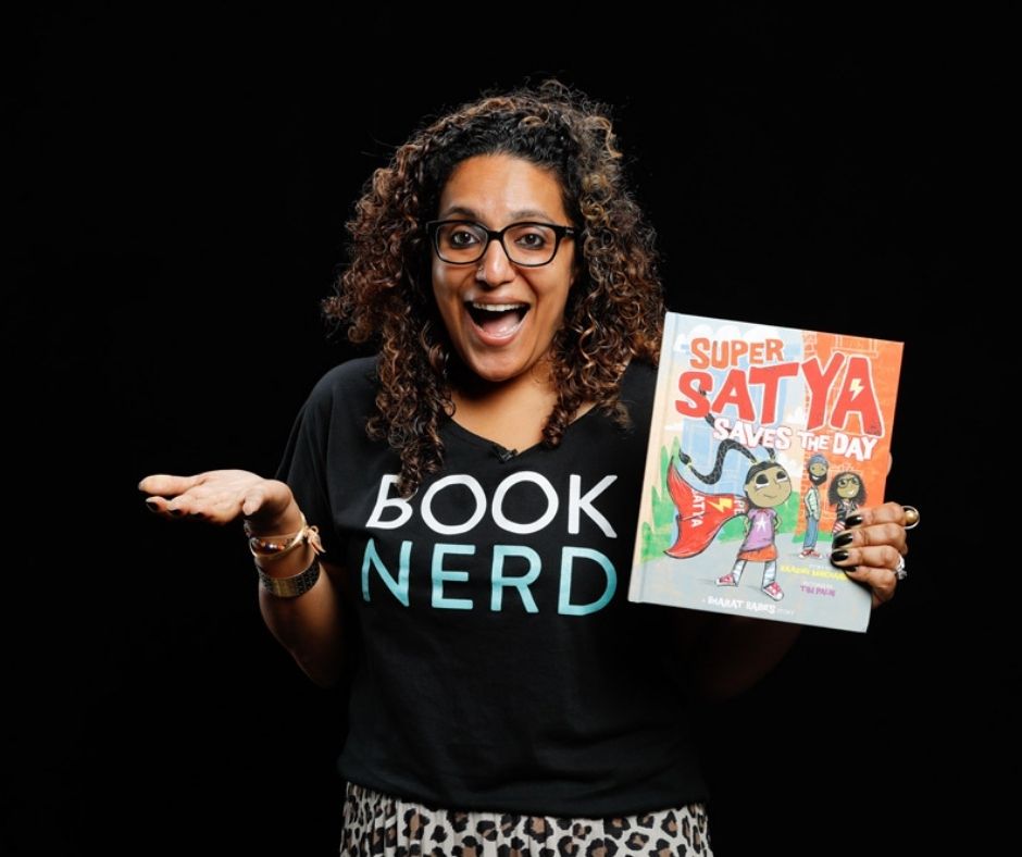 Image of Raakhee Mirchandani holding her book Super Satya. she is has a tee shirt that reads 'book nerd' and long curly hair, and glasses